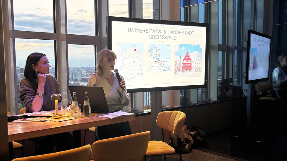 Zwei Frauen stehen an einem Tisch mit Laptop und Getränken. Eine von ihnen hält ein Mikrofon, hinter ihnen ist ein Bildschirm. (Quelle: Bundesamt für Migration und Flüchtlinge) Zwei Frauen stehen an einem Tisch mit Laptop und Getränken. Eine von ihnen hält ein Mikrofon, hinter ihnen ist ein Bildschirm.