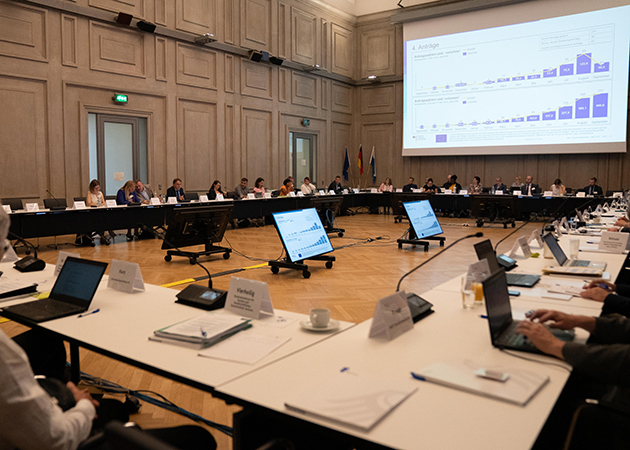 Blick in einen großen Konferenzsaal, in dem mehrere Personen an Tischen sitzen. Im Hintergrund wird eine Präsentation auf eine Leinwand projeziert. (Quelle: BAMF) Blick in einen großen Konferenzsaal, in dem mehrere Personen an Tischen sitzen. Im Hintergrund wird eine Präsentation auf eine Leinwand projeziert.