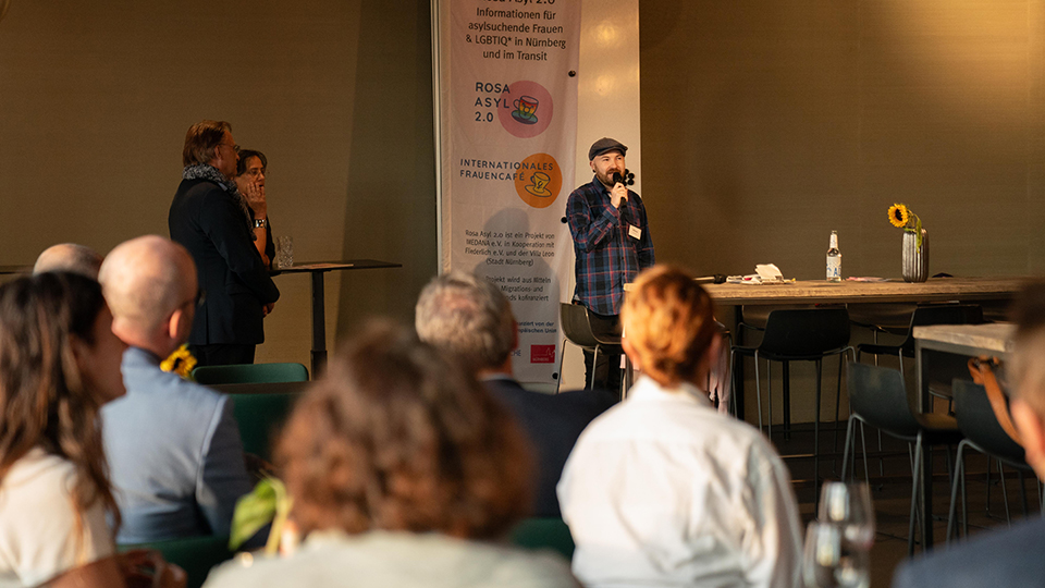 Ein Mann steht in einem Raum und spricht in ein Mikrofon. Mehrere Personen sitzen vor ihm und hören ihm zu. (Quelle: BAMF) Ein Mann steht in einem Raum und spricht in ein Mikrofon. Mehrere Personen sitzen vor ihm und hören ihm zu.