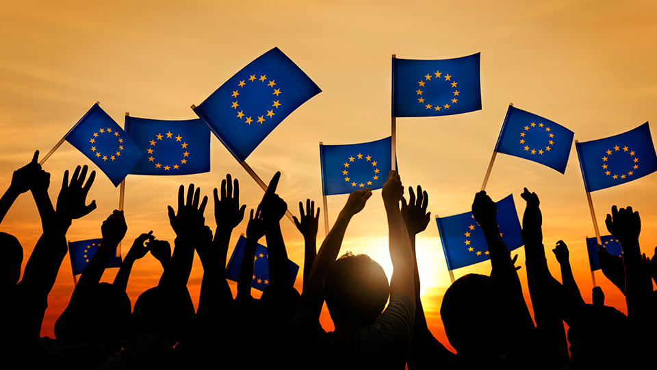 Eine Gruppe von Menschen winkt, während EU-Flaggen im Gegenlicht wehen. Im Hintergrund ist eine helle Sonne zu sehen, wodurch die Silhouetten der Personen sichtbar werden. (Quelle: iStock | Rawpixel) Eine Gruppe von Menschen winkt, während EU-Flaggen im Gegenlicht wehen. Im Hintergrund ist eine helle Sonne zu sehen, wodurch die Silhouetten der Personen sichtbar werden.