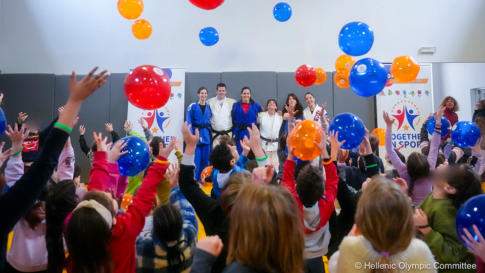 Frauen und Männer stehen auf einer Bühne. Davor stehen viele Menschen, die Luftballons in die Luft schmeißen.  (Quelle: METAdrasi / © Hellenic Olympic Committee) Frauen und Männer stehen auf einer Bühne. Davor stehen viele Menschen, die Luftballons in die Luft schmeißen.