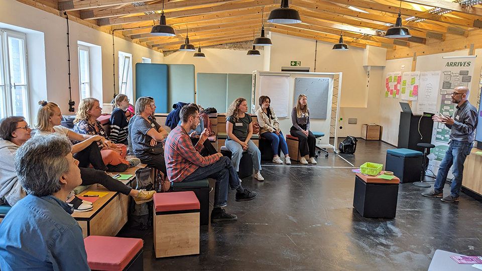 Ein Mann, stehend, hält eine Rede vor einer Gruppe von Frauen und Männern, die auf Hockern sitzen und aufmerksam zuhören. (Quelle: Social Impact gGmbH) Ein Mann, stehend, hält eine Rede vor einer Gruppe von Frauen und Männern, die auf Hockern sitzen und aufmerksam zuhören.