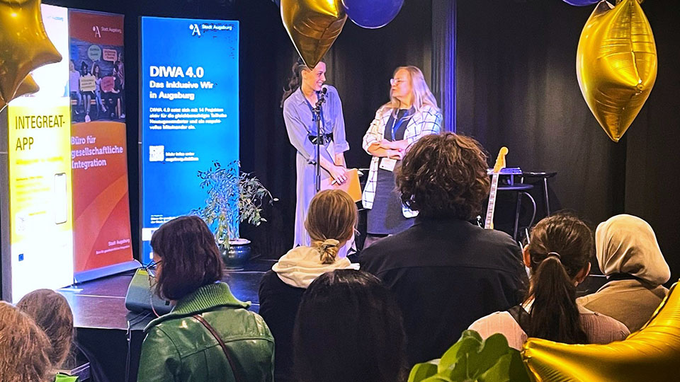 Zwei Frauen stehen auf einer Bühne und halten vor Publikum eine Rede. Der Raum ist mit Luftballons und Werbeplakaten dekoriert. (Quelle: Stadt Augsburg) Zwei Frauen stehen auf einer Bühne und halten vor Publikum eine Rede. Der Raum ist mit Luftballons und Werbeplakaten dekoriert.