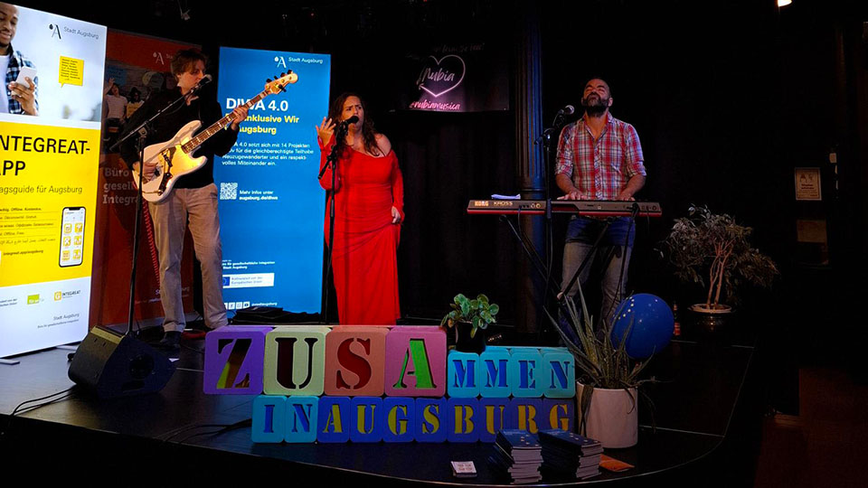 Auf einer Bühne steht eine Frau in einem roten Kleid. Sie macht gemeinsam mit zwei Männern Musik – einer spielt Gitarre, der andere Keyboard. (Quelle: Stadt Augsburg) Auf einer Bühne steht eine Frau in einem roten Kleid. Sie macht gemeinsam mit zwei Männern Musik – einer spielt Gitarre, der andere Keyboard.