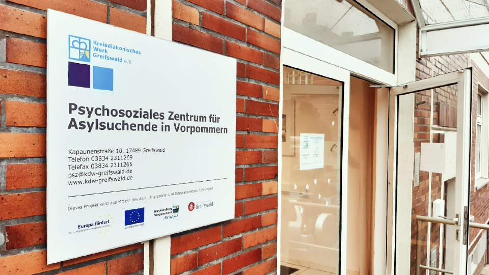 Eingangsbereich eines Gebäudes mit offener Tür und Schild an der Wand, auf dem Adresse, Kontaktdaten und Logo des Psychosozialen Zentrums für Asylsuchende in Vorpommern sowie das EU-Emplem abgebildet sind (Quelle: Kreisdiakonisches Werk Greifswald e.V.) Eingangsbereich eines Gebäudes mit offener Tür und Schild an der Wand, auf dem Adresse, Kontaktdaten und Logo des Psychosozialen Zentrums für Asylsuchende in Vorpommern sowie das EU-Emplem abgebildet sind