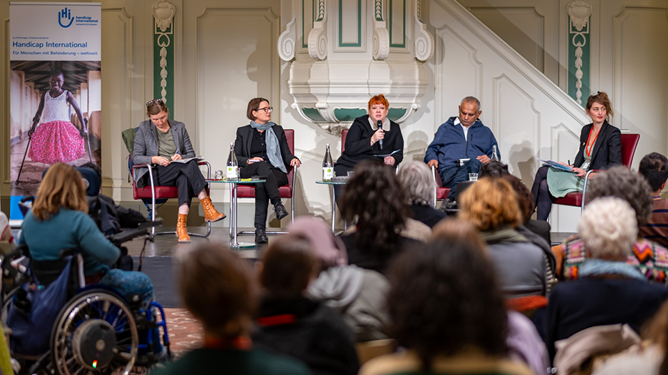 Vier Frauen und ein Mann sitzen auf einer Bühne vor Publikum. Hinter ihnen steht ein Aufsteller von "Handicap International", auf dem ein Mädchen mit Krücken abgebildet ist. (Quelle: Handicap International e.V.) Vier Frauen und ein Mann sitzen auf einer Bühne vor Publikum. Hinter ihnen steht ein Aufsteller von "Handicap International", auf dem ein Mädchen mit Krücken abgebildet ist.