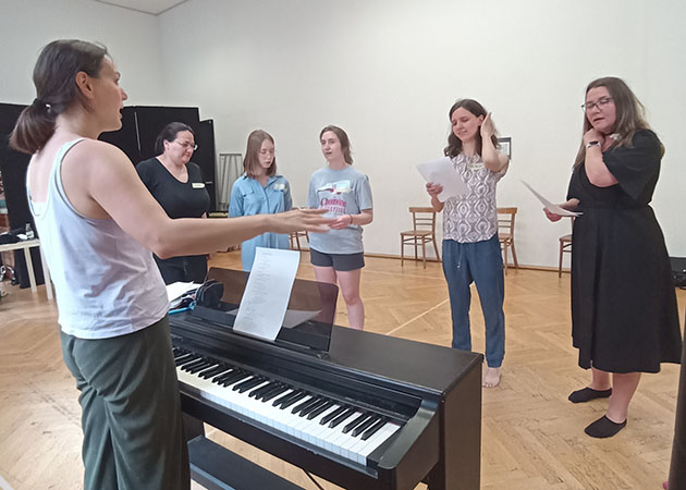 Eine Frau singt am Klavier, fünf weitere stehen ihr gegenüber, singen und halten Notenblätter in den Händen (Quelle: karl-arnold-stiftung.de) Eine Frau singt am Klavier, fünf weitere stehen ihr gegenüber, singen und halten Notenblätter in den Händen