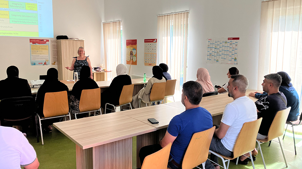 Mehrere Frauen und Männer sitzen in zwei Reihen in einem Klassenzimmer. Eine Lehrkraft spricht, während ein Beamer Inhalte an die Wand projiziert. (Quelle: backontrackev.org) Mehrere Frauen und Männer sitzen in zwei Reihen in einem Klassenzimmer. Eine Lehrkraft spricht, während ein Beamer Inhalte an die Wand projiziert.