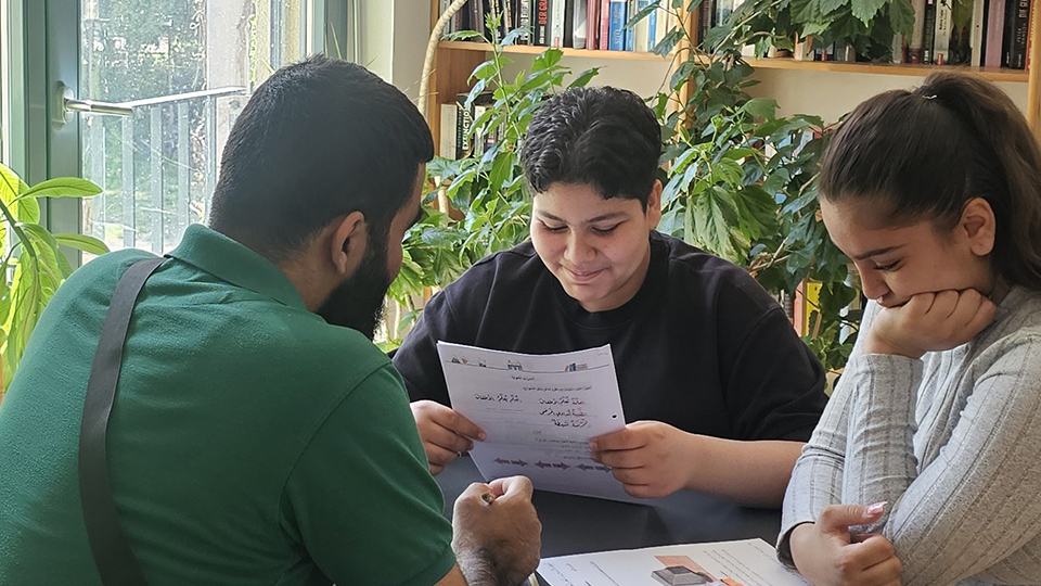 Ein Mädchen und ein Junge sitzen mit einem Lehrenden an einem Tisch. Sie halten Arbeitsblätter in der Hand. Im Hintergrund steht ein Bücherregal. (Quelle: backontrackev.org) Ein Mädchen und ein Junge sitzen mit einem Lehrenden an einem Tisch. Sie halten Arbeitsblätter in der Hand. Im Hintergrund steht ein Bücherregal.