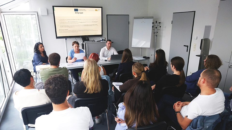 Zwei Frauen und ein Mann sitzen vor mehreren Teilnehmenden; im Hintergrund ist ein Bildschirm mit einer Präsentation zu sehen. (Quelle: Bayerisches Landesamt für Asyl und Rückführungen (LfAR)) Zwei Frauen und ein Mann sitzen vor mehreren Teilnehmenden; im Hintergrund ist ein Bildschirm mit einer Präsentation zu sehen.