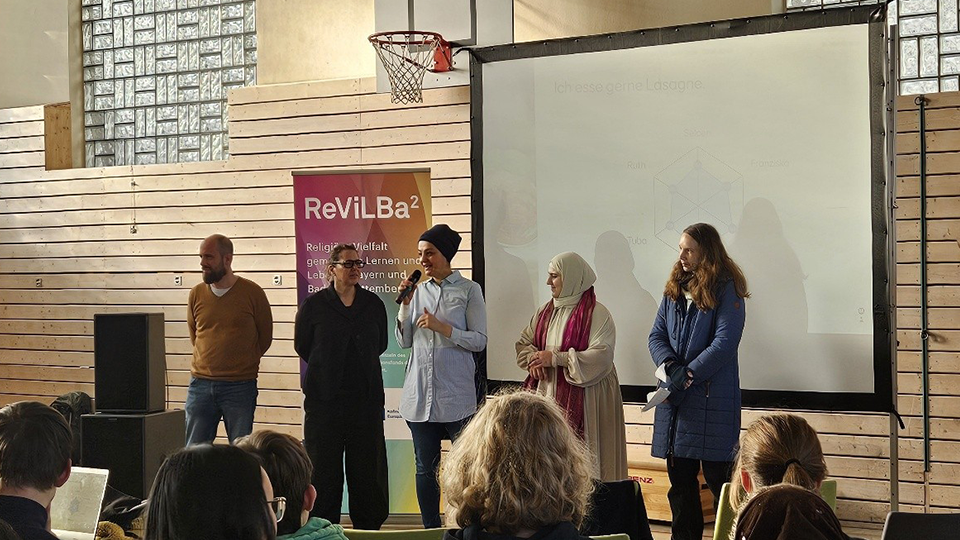 In einer Turnhalle stehen vier Frauen und ein Mann vor einer großen Leinwand. Eine der Frauen spricht mit einem Mikrofon zum Publikum. (Quelle: Eugen-Biser-Stiftung) In einer Turnhalle stehen vier Frauen und ein Mann vor einer großen Leinwand. Eine der Frauen spricht mit einem Mikrofon zum Publikum.