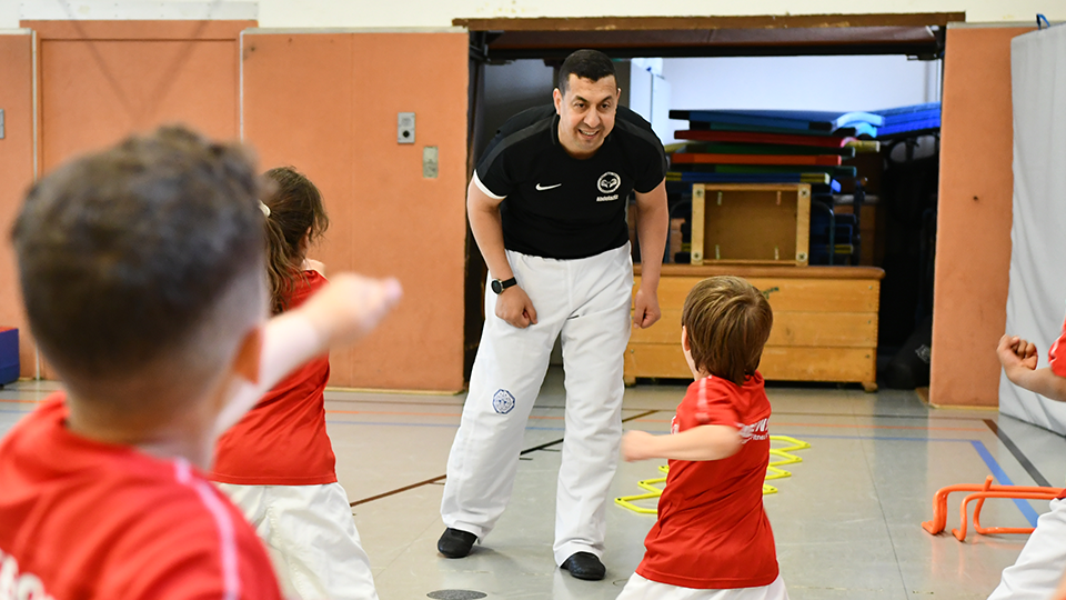 Mehrere Kinder führen in einer Turnhalle Taekwondo-Übungen aus. Vor ihnen steht lächelnd ihr Taekwondo-Trainer. (Quelle: BAMF | Kristin Kasten) Mehrere Kinder führen in einer Turnhalle Taekwondo-Übungen aus. Vor ihnen steht lächelnd ihr Taekwondo-Trainer.